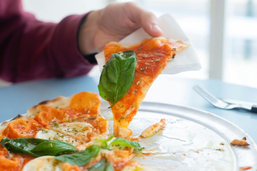 Close-up of a hand holding a slice of delicious Margherita pizza with basil topping on a sunny day.