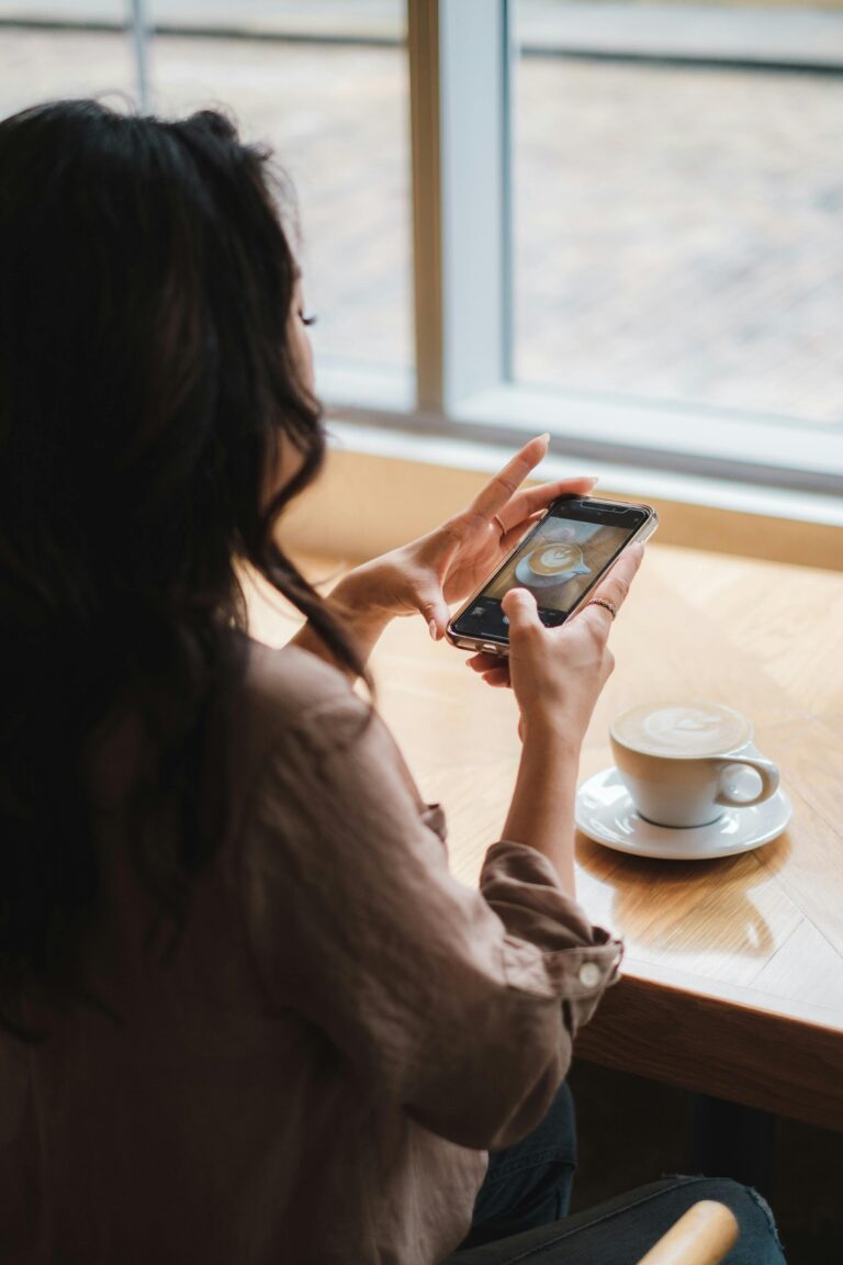 Rear view of woman capturing latte art on phone in a sunny cafe.